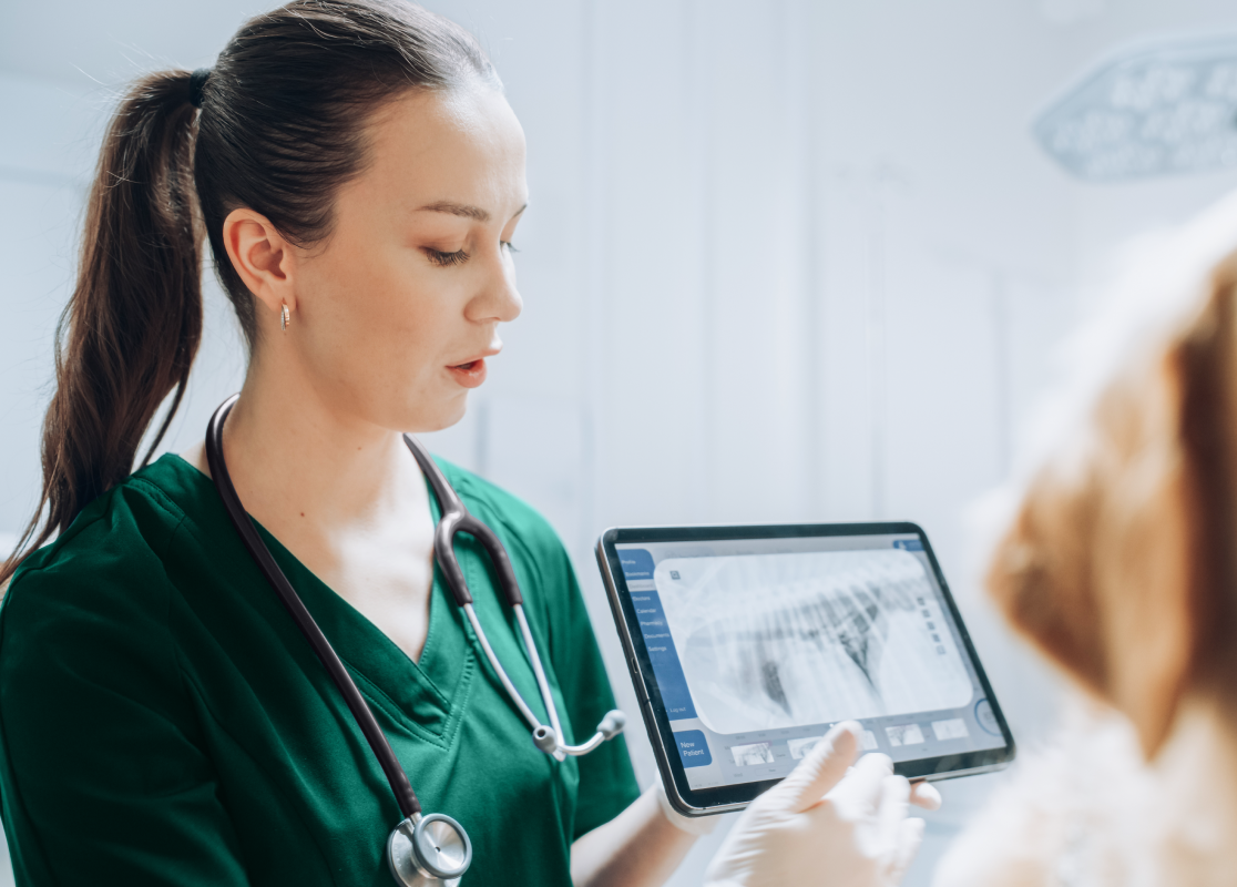 Eine Veterinärmedizinerin mit Stethoskop und grünem OP-Kittel (Antech) erklärt Röntgenaufnahmen auf einem Tablet. Im Vordergrund ist das verschwommene Gesicht eines Hundes zu sehen.